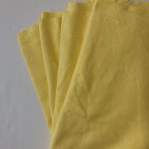 Pode incluir: Um leque de tecido amarelo claro dobrado. O material é liso e parece ser algodão ou um têxtil semelhante. As bordas do tecido são cuidadosamente acabadas, sugerindo um artigo costurado como uma toalha de mesa ou guardanapos.