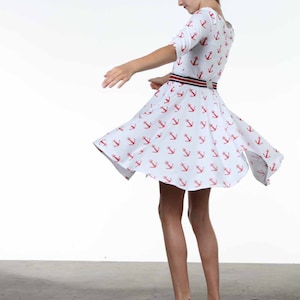 May include: A young person wearing a white dress with a red anchor print and a blue and white striped belt. They are wearing blue wedge sandals and are twirling.
