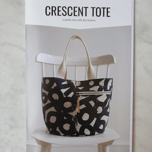 May include: A tote bag with a black and white abstract design, with a zipped pocket and cream-coloured handles. The bag is on a white chair, with the text "CRESCENT TOTE" and "noodlehead" visible.