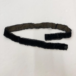 May include: A black velvet belt with a decorative trim and a button closure. The belt has a quilted pattern and a dark brown backing. The trim is a delicate, scalloped design. The belt is shown on a white background.