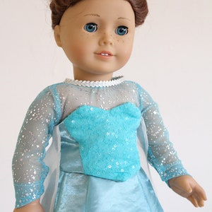 May include: A doll wearing a light blue dress with a sparkly bodice and a white cape. The doll has brown hair and blue eyes.