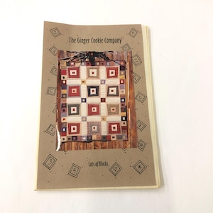 Puede incluir: Un folleto de patrones de papel marrón para una colcha titulada "Lots of Blocks" de The Ginger Cookie Company. La colcha presenta un diseño de patchwork con cuadrados de tela roja, blanca, azul y verde.