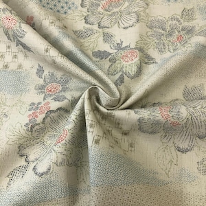 May include: A cream-colored fabric featuring a repeating pattern of large floral motifs in shades of blue, gray, and red. The design includes intricate geometric textures and grid-like details integrated into the background and petals.