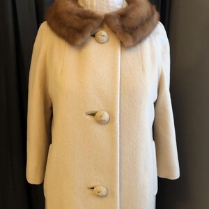 May include: A vintage cream-coloured coat with a brown fur collar. The coat has three large, decorative buttons and a classic, tailored design. The coat is displayed on a mannequin, showcasing its shape and style.