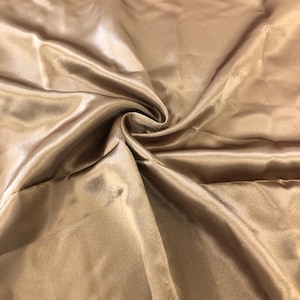 May include: A smooth, silky fabric in a light brown color. The fabric is folded and draped, showcasing its luxurious texture.
