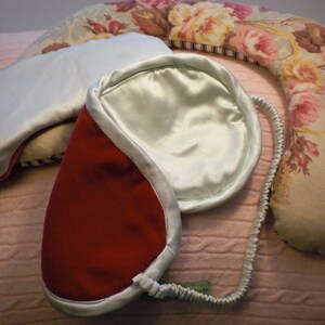 Può includere: Due maschere per dormire, una rossa e una blu chiaro, con un bordo in raso blu chiaro. Le maschere sono su una superficie a maglia rosa.