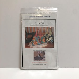 May include: A clear plastic package containing a sewing pattern for a Caddy Pad. The pattern includes instructions for a clever iron tote and ironing pad. The package features a photo of the finished product, a floral tote bag, and includes heat-resistant fabric.