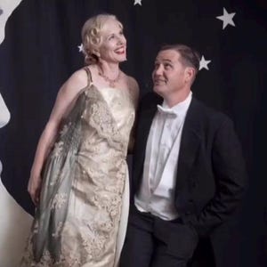 Puede incluir: Una pareja vestida con atuendo vintage posa frente a un fondo negro con decoraciones de luna creciente y estrellas. La mujer lleva un vestido color crema con detalles de encaje, el hombre un esmoquin.