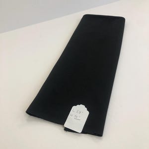May include: A folded piece of solid black fabric with a white rectangular tag attached. The tag features handwritten text that reads L 58" and W 34", indicating the dimensions of the material in inches. The fabric has a smooth, matte texture.