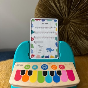 May include: A teal toy piano with a colourful keyboard and a card that says "Baby Shark" with musical notes and lyrics. The card is in a slot on the piano.