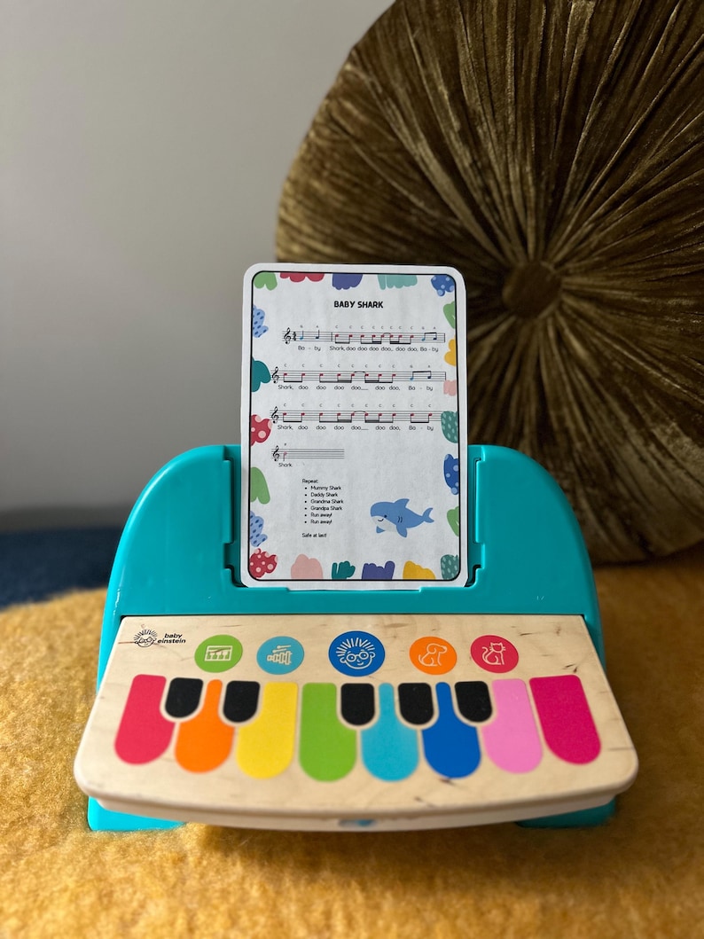 May include: A teal toy piano with a colourful keyboard and a card that says "Baby Shark" with musical notes and lyrics. The card is in a slot on the piano.