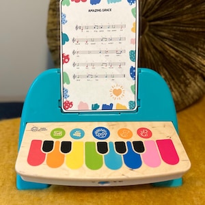 以下が含まれることがあります： カラフルな鍵盤と「Amazing Grace」と書かれた楽譜がついた青いおもちゃのピアノ。ピアノには楽譜を置くスタンドがついています。