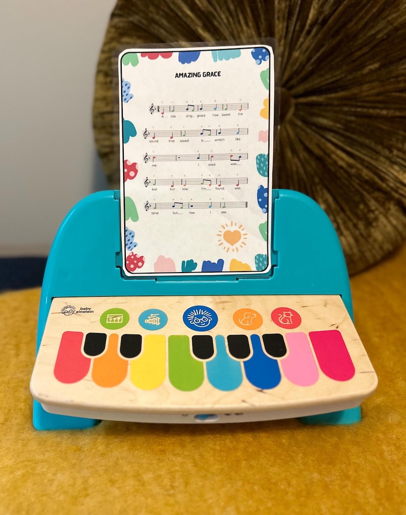 以下が含まれることがあります： カラフルな鍵盤と「Amazing Grace」と書かれた楽譜がついた青いおもちゃのピアノ。ピアノには楽譜を置くスタンドがついています。