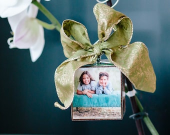 May include: A square photo ornament with a picture of two children, adorned with a green and gold bow. The ornament is suspended from a silver wire and is attached to a plant stem, creating a decorative display.