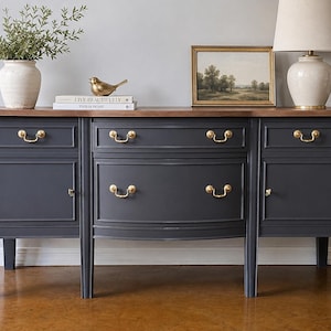 May include: A dark charcoal gray sideboard featuring a natural wood top, two side cabinets, and three central drawers. The furniture piece is accented with polished brass hardware, including curved drawer pulls and small round cabinet knobs.