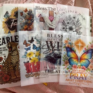 May include: A collection of translucent vellum sheets featuring various religious and inspirational designs, including butterflies, flowers, a leopard, a cross, and text such as Bless The Lord, Fearless, and Pray About Everything.
