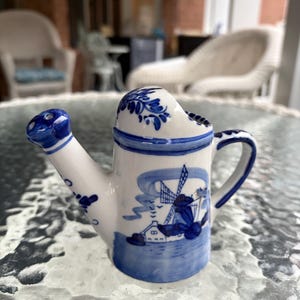 May include: A small, white ceramic watering can with blue floral and windmill designs. The handle and spout are blue, and the can sits on a glass table. The overall design is reminiscent of Delftware.