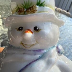 May include: A glossy white snowman figurine with a top hat decorated with pine needles and pine cones. The snowman has a carrot nose, black eyes, and a purple and blue scarf. The snowman has a smiling expression.