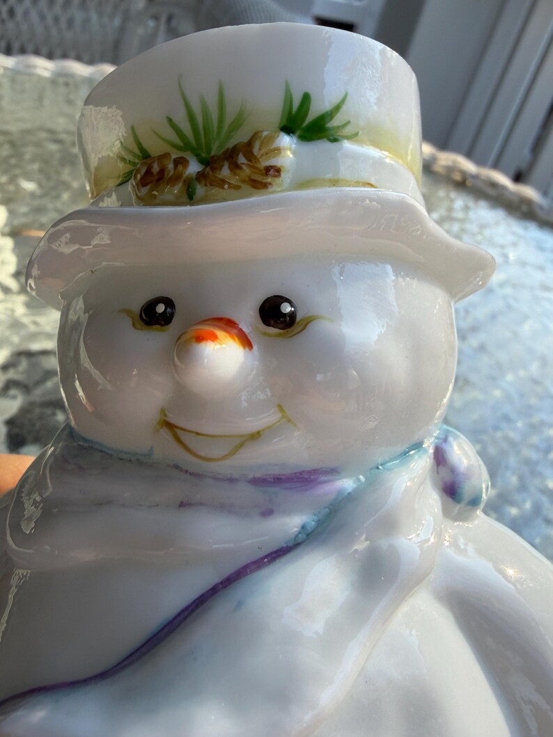 May include: A glossy white snowman figurine with a top hat decorated with pine needles and pine cones. The snowman has a carrot nose, black eyes, and a purple and blue scarf. The snowman has a smiling expression.