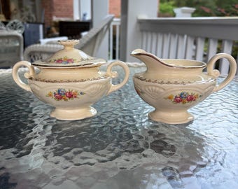 May include: A cream-colored sugar bowl with a lid and a creamer set. Both pieces have floral designs in pink, blue, and yellow, with gold trim. The set is placed on a glass table, suggesting a vintage or antique style.