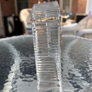K&ouml;nnte beinhalten: Eine klare, rechteckige Kristallflasche mit horizontalen Rillen. Die Flasche hat ein goldfarbenes Band um den oberen Rand. Die Flasche steht auf einem Glastisch.