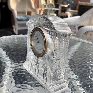 K&ouml;nnte beinhalten: Eine klare Kristalluhr mit goldfarbenem Rahmen und wei&szlig;em Zifferblatt. Die Uhr hat eine rechteckige Form mit einem dekorativen, strukturierten Design. Die Uhr steht auf einem Glastisch.