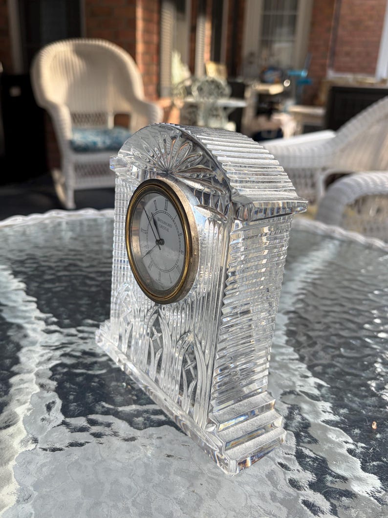 K&ouml;nnte beinhalten: Eine klare Kristalluhr mit goldfarbenem Rahmen und wei&szlig;em Zifferblatt. Die Uhr hat eine rechteckige Form mit einem dekorativen, strukturierten Design. Die Uhr steht auf einem Glastisch.