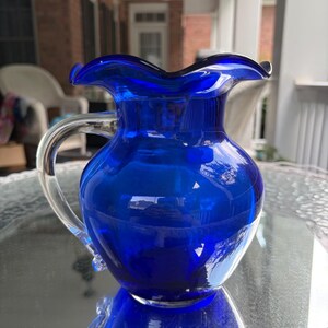 May include: A cobalt blue glass pitcher featuring a rounded body, a clear glass handle, and a ruffled, wavy rim. The pitcher has a smooth, glossy finish and a translucent appearance, reflecting light across its curved surface.