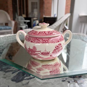 May include: A cream-colored sugar bowl with a red and white landscape design. The bowl has a lid with a small knob and two handles. The design features a boat, trees, and buildings. The sugar bowl is sitting on a glass table.