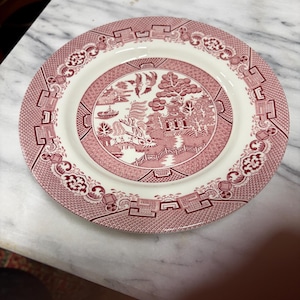 May include: A round, white and red decorative plate with a detailed landscape design in the center. The plate has a red border with repeating geometric and floral patterns. The plate is made of ceramic.