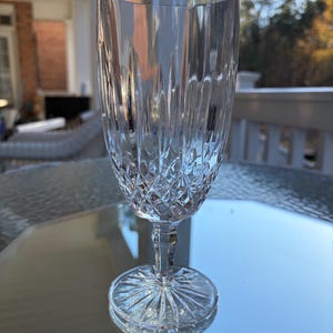 May include: A clear crystal champagne flute with a diamond-cut pattern on the bowl and a starburst design on the base. The stem is slender, and the glass reflects light, creating a sparkling effect. The glass is on a reflective surface.