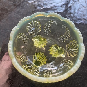 May include: A clear glass bowl with a scalloped edge and a pale yellow-green hue. The bowl features embossed floral and leaf designs in a lighter shade of green. The bowl is likely vintage or antique.