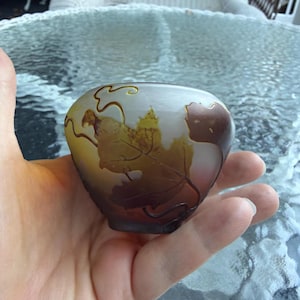 May include: A small, oval-shaped art glass vase with a leaf design. The vase features a gradient of colors, transitioning from a light yellow to a deep brown. The leaf pattern is etched into the glass, creating a textured effect. The vase is held in a hand.