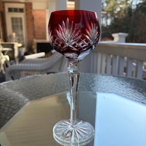 May include: A ruby red crystal wine glass with a clear, long stem and a circular base. The bowl features a cut crystal design. The glass is displayed on a reflective surface, with a blurred outdoor background.