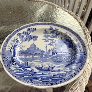 May include: A white and blue decorative plate with a scenic landscape design. The plate features a detailed illustration of a bridge, buildings, trees, and boats. The rim of the plate has a decorative border.
