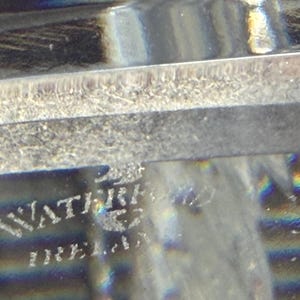 K&ouml;nnte beinhalten: Nahaufnahme eines transparenten Glasobjekts mit dem eingravierten Text "WATERFORD IRELAND". Das Glas hat eine strukturierte Oberfl&auml;che und reflektiert das Licht, wodurch ein Regenbogeneffekt entsteht. Das Objekt scheint ein dekoratives St&uuml;ck zu sein.