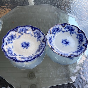 May include: Two round ceramic bowls featuring a white base with a blue floral pattern and scalloped edges. The rims are accented with gold trim, and the interior surfaces display a central blue flower motif surrounded by intricate blue floral detailing.
