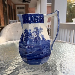 May include: A blue and white ceramic pitcher with a landscape design. The pitcher features a detailed illustration of a village scene with trees, buildings, and a bridge. The handle and rim are adorned with floral patterns.