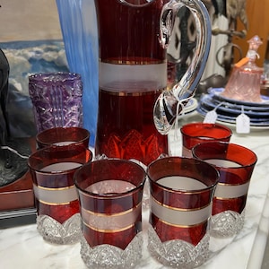May include: A vintage red glass pitcher and matching glasses set. The pitcher has a clear, curved handle and a frosted band with gold trim. The glasses have a similar design with a decorative, cut-glass base. The set is displayed on a marble surface.