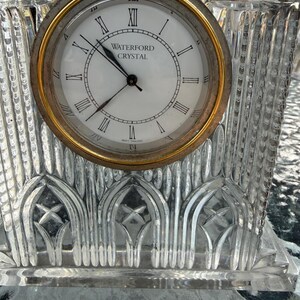 K&ouml;nnte beinhalten: Eine Uhr aus klarem Kristall mit goldfarbener L&uuml;nette und wei&szlig;em Zifferblatt. Das Zifferblatt hat schwarze r&ouml;mische Ziffern und die Worte "WATERFORD CRYSTAL". Die Uhr hat einen dekorativen, geschliffenen Kristallfu&szlig;.