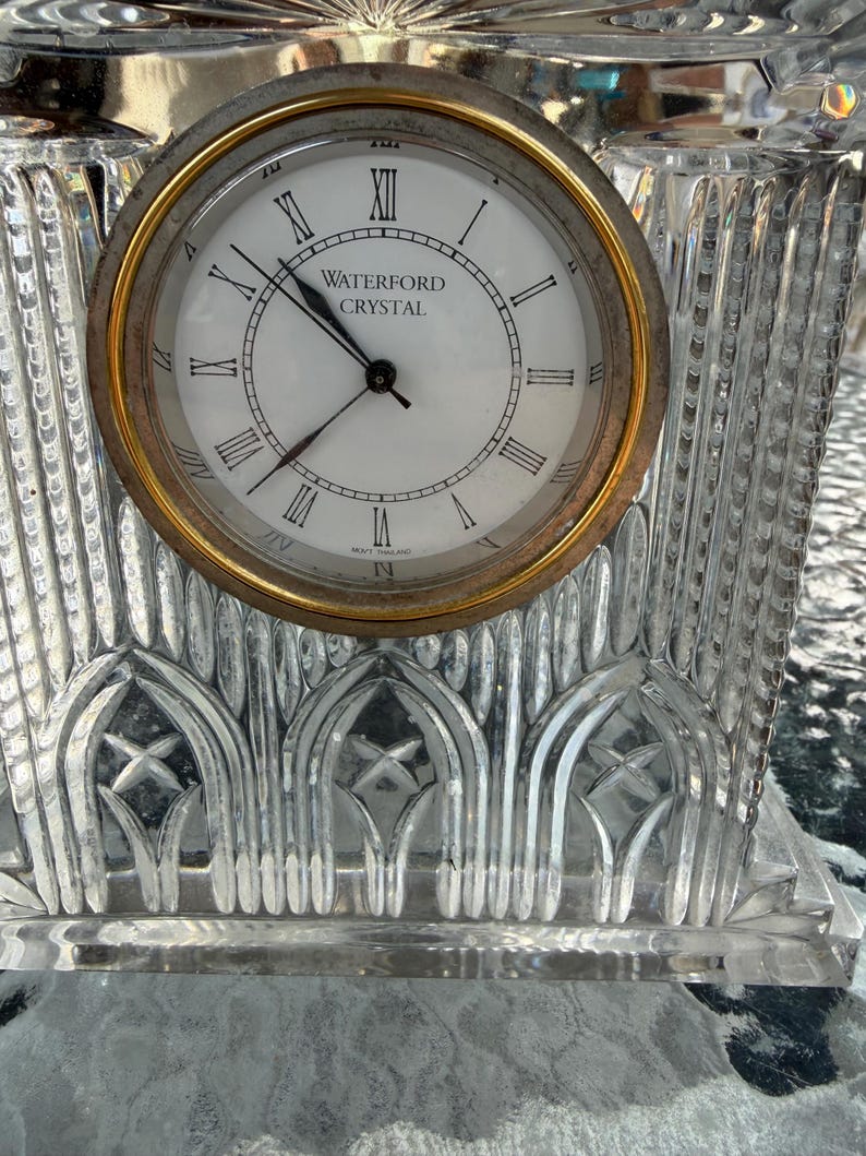 K&ouml;nnte beinhalten: Eine Uhr aus klarem Kristall mit goldfarbener L&uuml;nette und wei&szlig;em Zifferblatt. Das Zifferblatt hat schwarze r&ouml;mische Ziffern und die Worte "WATERFORD CRYSTAL". Die Uhr hat einen dekorativen, geschliffenen Kristallfu&szlig;.