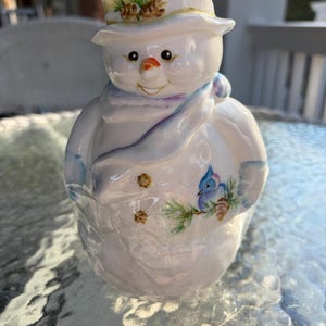 May include: A ceramic snowman figurine with a top hat, scarf, and painted details. The snowman has a carrot nose, coal eyes, and a painted bluebird on its side. The hat is decorated with pine needles and pine cones.