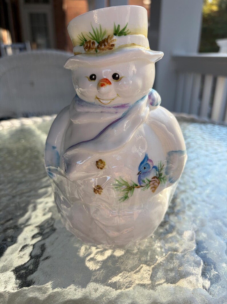 May include: A ceramic snowman figurine with a top hat, scarf, and painted details. The snowman has a carrot nose, coal eyes, and a painted bluebird on its side. The hat is decorated with pine needles and pine cones.