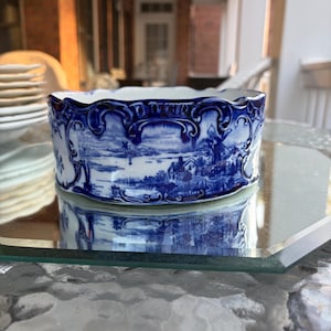 May include: A blue and white ceramic bowl with a landscape design. The bowl has a scalloped rim and is sitting on a glass surface. The bowl's design features a detailed depiction of a landscape scene.
