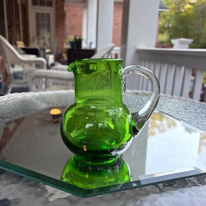 May include: A small, green glass pitcher with a rounded base and a clear handle. The pitcher is sitting on a glass surface, reflecting its shape. The pitcher has a spout for pouring.