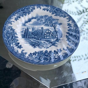 May include: A round ceramic plate featuring a blue and white transferware pattern. The center depicts a rural landscape with a cottage, trees, and a stream. The rim is decorated with a detailed floral and geometric border in blue and white.