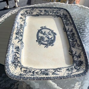 May include: Rectangular serving platter with a cream-colored base and a blue floral design. The border features a detailed pattern, and the center has a circular emblem with a bird and floral motif. The platter is likely made of ceramic.