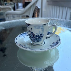 May include: A white teacup and saucer set with a blue windmill and floral design. The cup has a curved handle and sits on a matching saucer. The set is placed on a reflective surface.