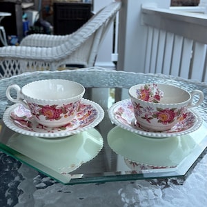 May include: Two white teacups and saucers with a floral pattern in pink, red, and yellow. The teacups have a scalloped rim and are placed on a mirrored surface. The saucers have a matching floral design and a beaded edge. The set is on a glass table.