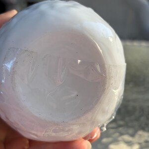 May include: A white, round ceramic object, possibly a bowl or cup, wrapped in clear plastic. The object is smooth and appears to be new, with a clean, pristine surface. The plastic wrap is secured with clear tape.